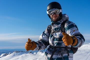 Smiling man in skiing or snowboarding winter equipment shows thumbs up. Warm jacket, brown gloves,...