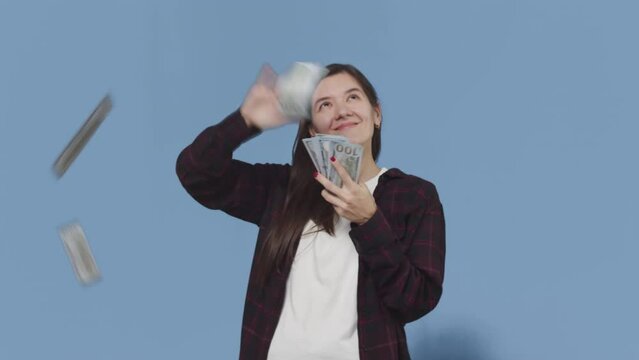 Young attractive woman with a smiling face throws banknotes around from the stack in all directions until it ends on a blue background. Concept of waste of money with pleasure