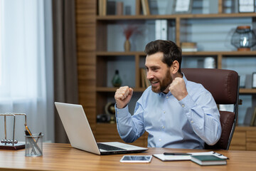 Exuberant mature businessman with a beard showing excitement and success at his home office desk, laptop screen visible.