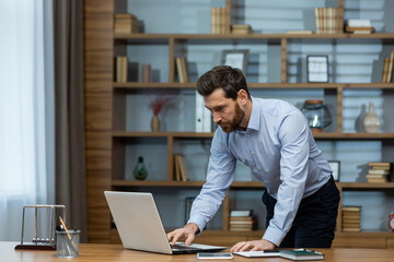 Obraz premium Focused mature businessman reviewing documents at his laptop in a well-organized, modern home office setting.
