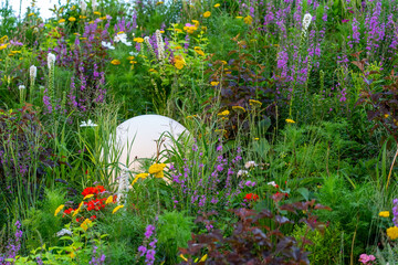 Naklejka premium White spherical lamp is surrounded by tall grass and wildflowers, including purple and pink plants. Scene is set during sunset, with warm, glowing colors.