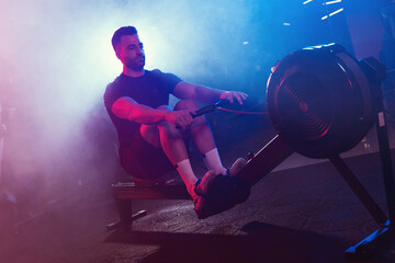 A male athlete pulls the oars on a rowing machine, his form lit by moody blue and red lights,...