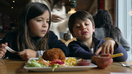 Children eating food at restaurant, siblings - small brother and sister enjoying fries and chicken meal at diner inside wooden interior