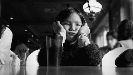 Depressed little girl feeling sad in monochromatic black and white at restaurant diner, child holds...