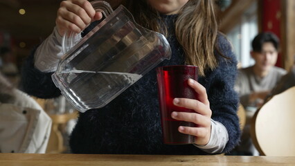 Little girl serving water into cup by holding jar inside restaurant, daughter giving cup to mother, tap water, caring gesture