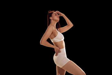 Fototapeta premium Slim, beautiful female model posing against black studio background. Woman wearing in sporty clothes, shorts and top. Concept of female health, fitness, sport, active lifestyle, dieting.
