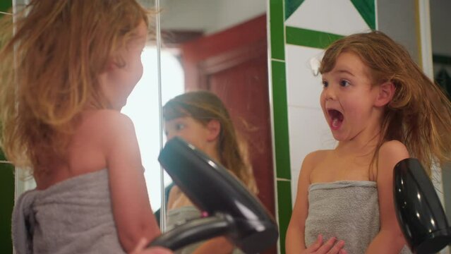 Cute little girl dries her hair with a hairdryer after a shower. The girl's hair is being dried while she looks in the mirror and makes faces
