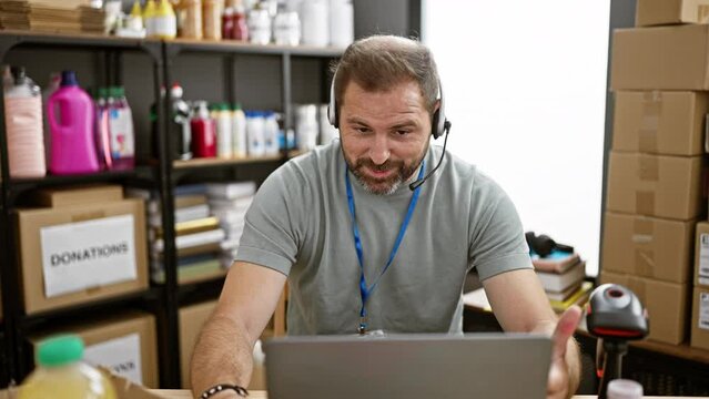 Smiling middle-aged man with grey hair having a video call at an indoor donation center, wearing a headset and lanyard.