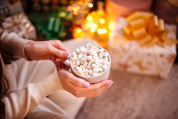 A white cup with cocoa and white marshmallows in female hands against a background of lights. Warm,...