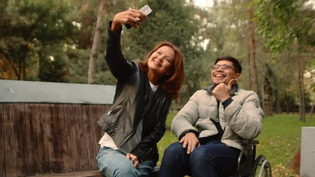 Disabled asian man in a wheelchair smiling and showing gestures in funny joint selfies with an asian woman on a smartphone in an autumn city park