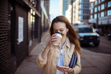 Happy woman with coffee and smartphone walking on sidewalk