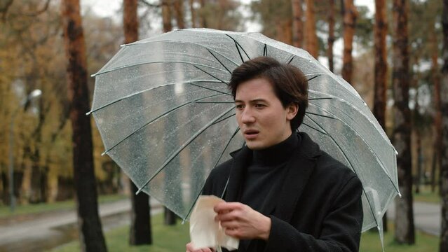 Young man in a coat standing under a transparent umbrella in an autumn city park and blowing his nose in a handkerchief because of wet and cold weather