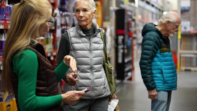 Mature woman with elderly man and woman trying to find and compare products in hardware store using smart phone in slow motion.