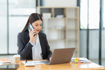 Shot of a young professional woman sitting at desk in front of laptop and using mobile phone working on business contract.