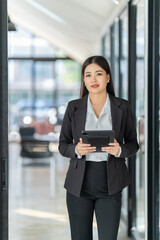 Portrait of beautiful young woman standing in front of glass board and smiling on camera. Female office worker using laptop in company.