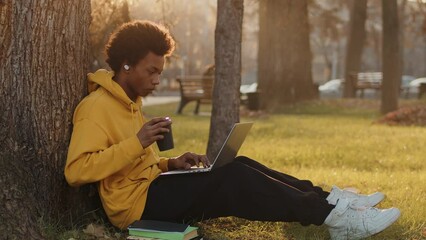 Young black programmer in a yellow hoodie sitting under a tree outdoors and writing program code on a laptop in a city park on a sunny day drinking coffee from a paper cup - Powered by Adobe