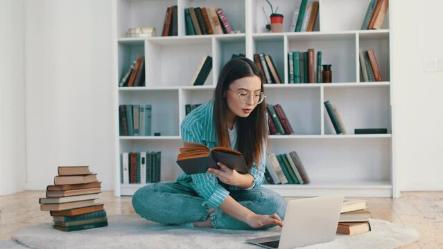 Young attractive woman doing education, sitting on the floor, flipping the pages of the book in her hands and looking at the laptop at home surrounded by stacks of books
