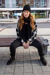 Woman in her mid-thirties with long red hair wearing a cap and black clothing in an urban location.