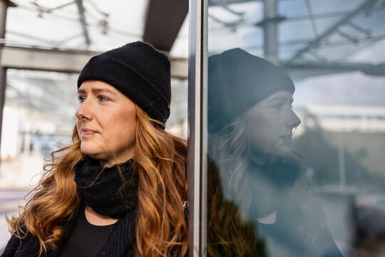 Woman in her mid-thirties with long red hair wearing a cap and black clothing in an urban location.