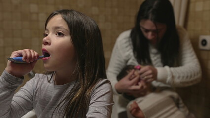 Child brushes teeth in front of bathroom mirror while mother assists smaller sibling to with dental...