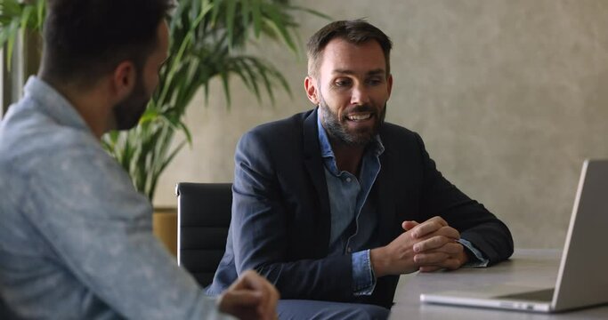 Mature businessman talking to male colleague sit at office table, speaking, brainstorming, explaining work tasks or new project details. Manager provide consultation to client during formal meeting