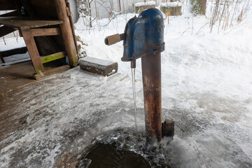 An old outdoor cast iron water heater in winter. A stream of water flows, turning into ice. There...