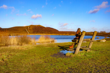 Autumn landscape with lake and two-level wooden bench with senior woman next to her dog taking a...