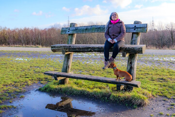 Two-level wooden bench with a senior adult woman next to her dog taking a break, looking up, path and bare trees in background, Thor Park - Hoge Kempen National Park, cloudy day in Genk, Belgium