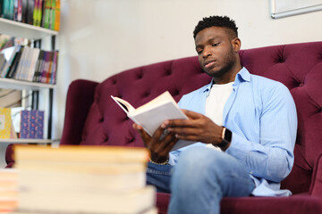 Serious young man engrossed in reading, surrounded by books at home, embracing the joy of learning.