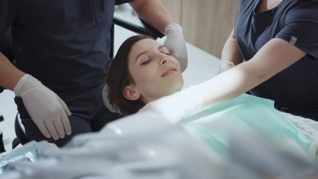 Shot of female Caucasian patient lying on dental chair. Nurse in blue uniform putting dental bib on woman in clinic. Healthcare concept. Stomatology. Indoors