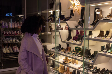 Fototapeta na wymiar The image presents a side view of a woman as she gazes into a shoe store window during an evening in the city. The well-lit display showcases a variety of shoes, from practical to high fashion