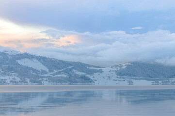 Fototapeta na wymiar A tranquil scene captures the serene beauty of a snow-covered landscape and a calm fjord under a soft winter sunset with fog lifting off the waters surface. 