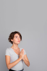 Fototapeta premium Vertical photo of cute young woman stands in meditative pose, enjoys peaceful atmosphere, hands in praying gesture and asking help, isolated over gray background, peace in soul