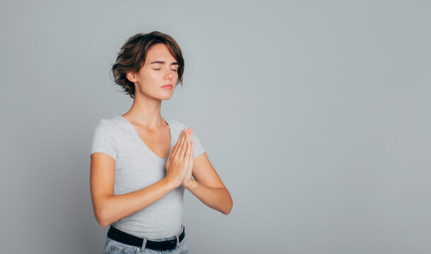 Peaceful young girl with closed eyes stands in meditative pose, enjoys peaceful atmosphere, hands in praying gesture and asking help, isolated over gray background, peace in soul