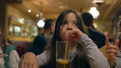 Pensive Little Girl Drinking Ice Tea with Straw at Restaurant in the Evening - Thoughtful Child Sipping Drink at Diner