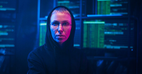 Caucasian female hacker in dark hood and glasses turning to camera on spinning chair and looking straight at night in room with monitors and data. Portrait of woman sitting in cyber police office.
