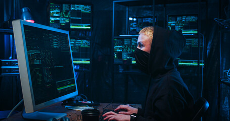 Side view Anonymous female Caucasian hacker with masked face working on computer in front of big...