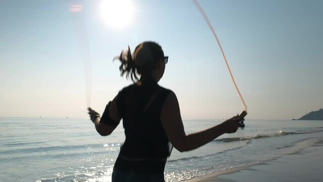 Silhouette of sporty woman working out on skipping rope on a sandy beach, slow motion. Active summer vacation on the sea and healthy lifestyle concept