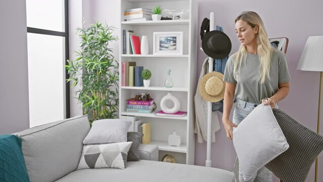 A young blonde woman arranges pillows on a couch in a modern living room with bookshelves and decor.