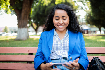 Young hispanic woman businesswoman using phone sitting on public park bench waiting for bus after...