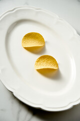 Two crispy potato chips on white vintage serving plate. White marble background. Minimalistic food photo. Concept of diet and Conscious eating 