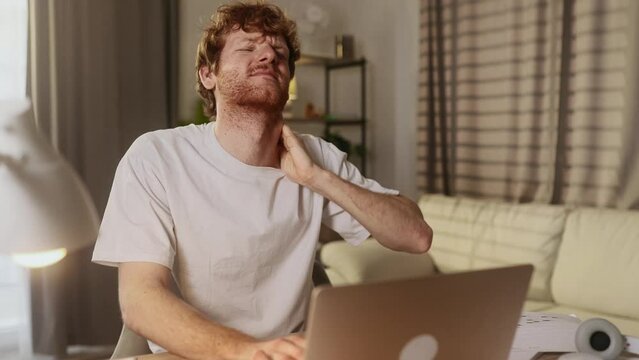 Overworked tired man freelancer or student with severe neck shoulder pain after hard work on laptop computer at home workplace Sad red haired male suffers from tension in the spine indoors