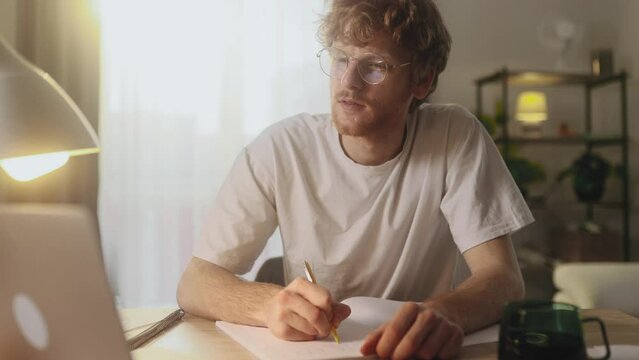 Handsome focused young man preparing for exam at university studying at home workplace Busy student make notes while looking at laptop computer screen and looking at camera Distance remote education	