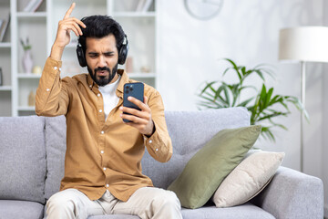 Happy young Indian man sitting at home on sofa wearing headphones and using mobile phone, relaxing,...