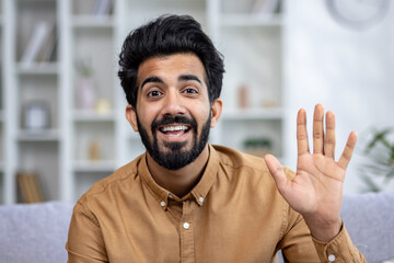 Close-up photo of a young Indian man sitting on the couch at home and chatting online. Says hello and waves at the camera. © Liubomir