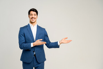 Young male business professional in a stylish blue suit pointing aside, his open hand suggesting a presentation or a welcoming introduction, set against a plain background - obrazy, fototapety, plakaty