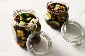Preserved and marinated cucumbers in jars in asian or Chinese style with soy sauce, garlic. Also includes ginger, chilli and other spices. White marble background