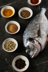 Raw and fresh dorado fish on black marble background with spices for cooking around. Minimalistic, dark-style or low key food photo. Mediterranean diet. 