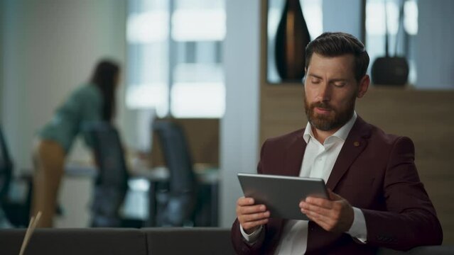 Sad millioner holding tablet computer in comfortable office interior cabinet.