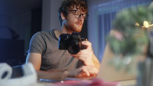 Handsome red haired young man photographer hold digital camera looking at screen choosing photos for editing sitting in front of computer at home workplace at late evening